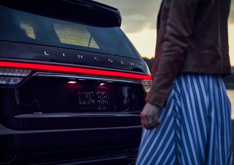 A person is shown near the rear of a 2024 Lincoln Aviator® SUV as the Lincoln Embrace illuminates the rear lights | Zeigler Lincoln of Kalamazoo in Kalamazoo MI