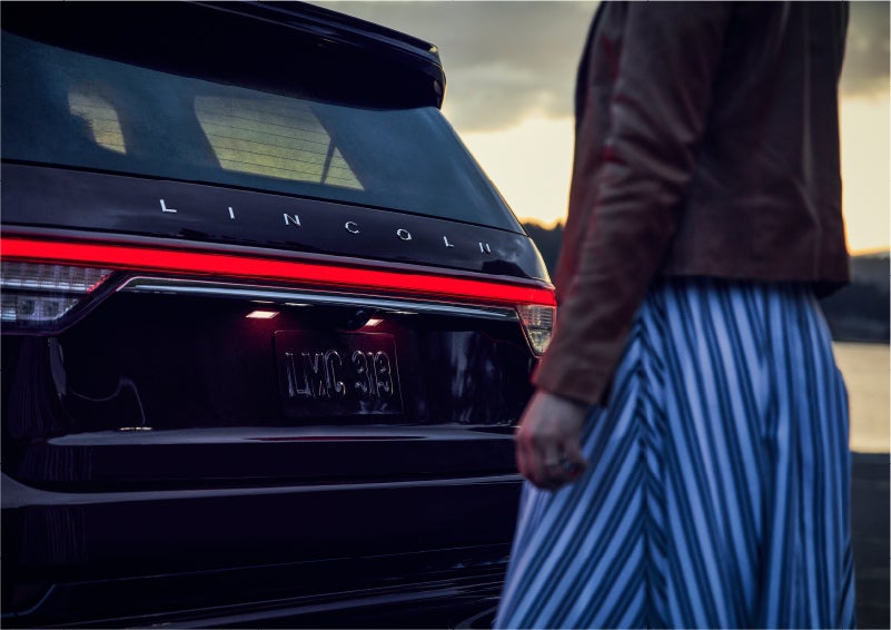 A person is shown near the rear of a 2023 Lincoln Aviator® SUV as the Lincoln Embrace illuminates the rear lights | Zeigler Lincoln of Kalamazoo in Kalamazoo MI